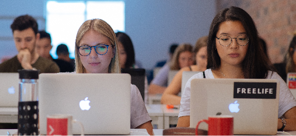 Two women with glasses sit at the front of a class on their laptops.